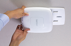 Ein Paar Hände installieren einen weißen, rechteckigen Access Point-Sockel mit abgerundeten Kanten an einer Decke. Die Basis verfügt über Laschen zum Anbringen des Access Point-Geräts.