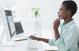 Person sitting at a desk in an office, facing a computer monitor with one hand on the mouse and the other near the chin in a contemplative gesture, looking into a dashboard.