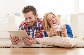 Zwei Personen liegen auf einem Teppichboden in einem Wohnzimmer. Eine Person hält ein Tablet in der Hand, die andere ein Smartphone. Sie ruhen beide auf dem Bauch, den Kopf auf die Hände gestützt. Der Hintergrund zeigt eine weiße Couch und einige Haushaltsgegenstände, die eine ungezwungene und entspannte Atmosphäre suggerieren, die möglicherweise auf Freizeit oder gemeinsame Aktivitäten mit digitalen Geräten hinweist.