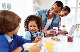 Eine Familie versammelt sich um eine Küchentheke. Zwei Kinder sitzen und frühstücken aus Schüsseln, während ein Erwachsener mit einer Tasse in der Hand daneben steht. Im Hintergrund arbeitet ein anderer Erwachsener an einem Laptop. An der Theke gibt es verschiedene Frühstücksartikel, darunter ein Glas Orangensaft und ein Glas Marmelade. Die Szene deutet auf eine geschäftige Morgenroutine in einer häuslichen Umgebung hin.