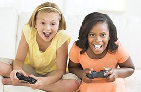 Zwei Personen sitzen auf einer Couch und halten jeweils einen Videospiel-Controller in der Hand. Die Person auf der linken Seite trägt ein gelbes Oberteil und beige Shorts, während die Person auf der rechten Seite ein orangefarbenes Oberteil und eine helle Hose trägt. Beide scheinen sich auf das Spiel zu konzentrieren, das sie spielen. Der Hintergrund zeigt eine helle Wand und einen Teil eines Fensters, was darauf hindeutet, dass es sich um ein Wohnzimmer handelt.