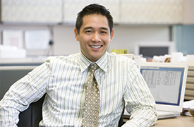 An individual sits in an office environment, wearing a striped shirt and a patterned tie. In the background, there are computer monitors, office supplies, and cubicle partitions, suggesting a typical corporate workspace setting.