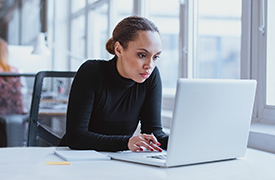 An individual sitting at a desk using a laptop, with their face obscured for privacy. Papers are on the desk, and another person is in the background, suggesting a work environment.