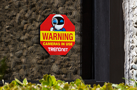 A red and yellow octagonal warning decal sign on a window. The sign features an illustration of a robot holding a magnifying glass, with the text “WARNING Cameras in use TRENDnet” below.
