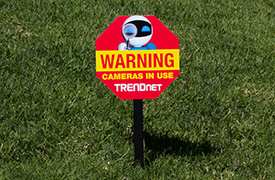 A red and yellow octagonal warning sign on green grass. The sign shows a robot with a magnifying glass and the text ‘WARNING Cameras in use TRENDnet’ below.