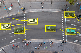 Vue aérienne d’une intersection routière à plusieurs voies avec plusieurs véhicules surlignés dans des cases jaunes, certains suivant des trajectoires courbes indiquées par des lignes jaunes. Les piétons sont visibles sur les trottoirs et traversant la rue. Cette scène se concentre sur la surveillance des véhicules et l’analyse du trafic.